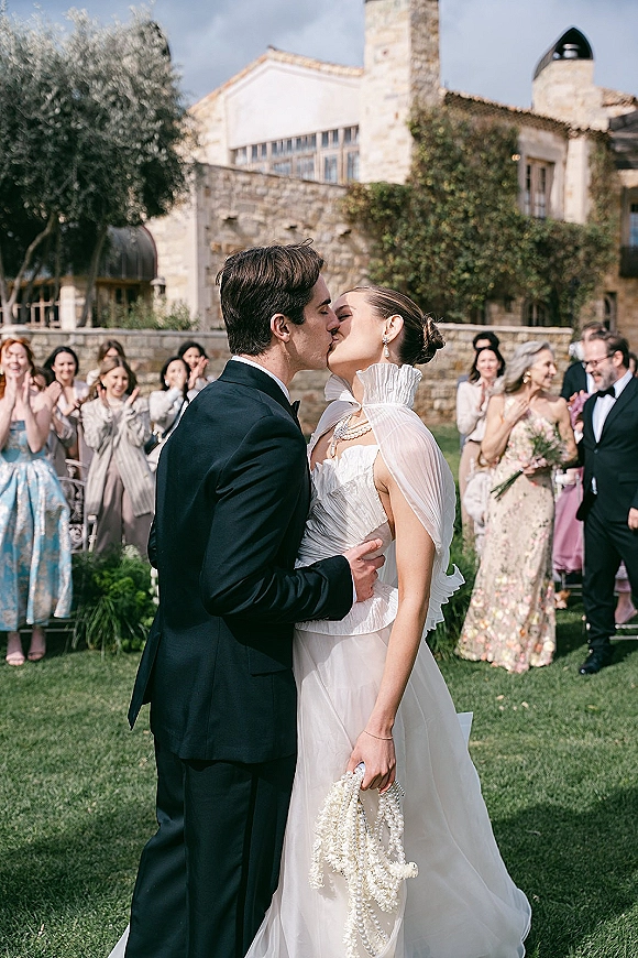 Wedding kiss as the bride in a veil and pearls holds a bouquet while guests cheer outside a stone building on a green lawn