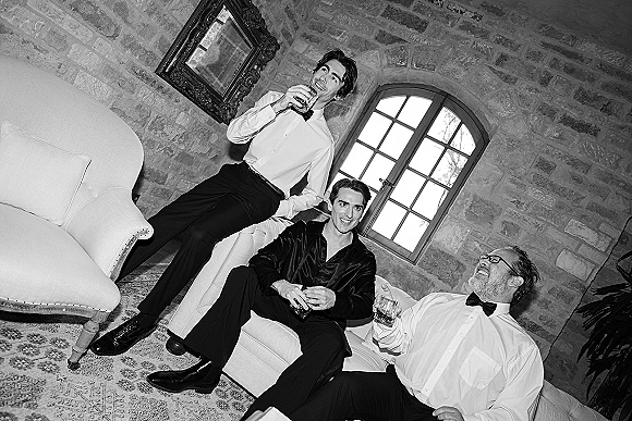 Groomsmen portrait of men in bow ties and white dress shirts holding whiskey glasses on a sofa in a rustic lounge with stone wall