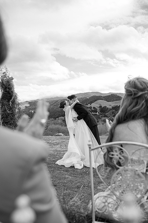 Wedding kiss portrait of bride and groom kissing beneath a floral arch, veil flowing, with mountain landscape and cloudy sky behind