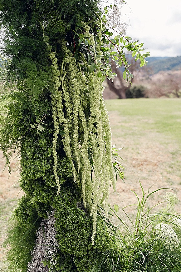 Ceremony floral arch with a green wedding arch draped in lush greenery and hanging amaranthus on a lawn with hills and trees under clouds