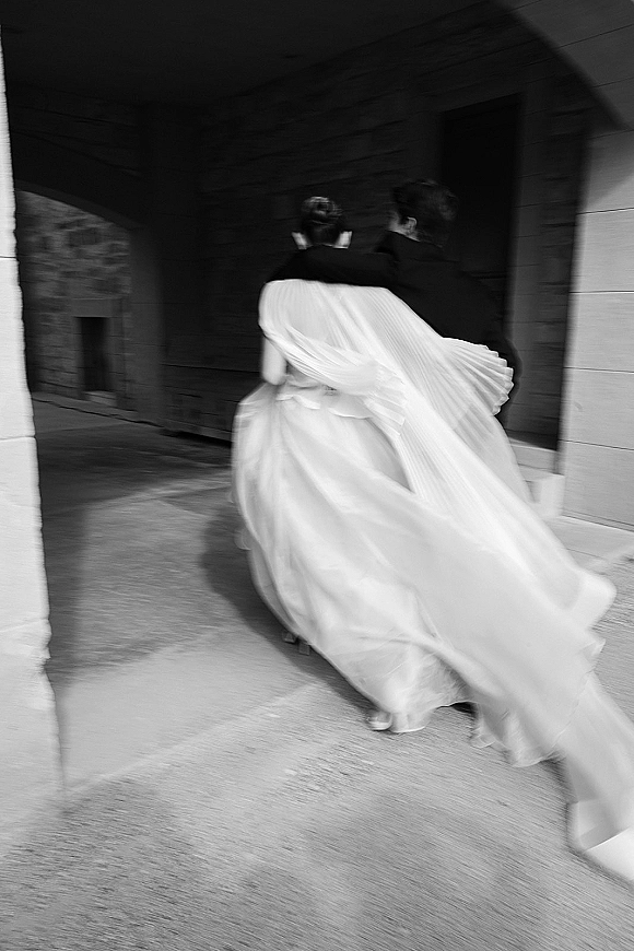 Wedding exit moment as bride and groom walk away under a stone archway, her long veil blowing in the wind behind her dress