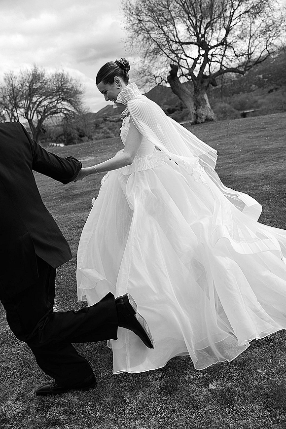 Couple portrait of bride and groom walking hand in hand as she twirls a cape veil wedding dress on a grassy lawn under cloudy hills