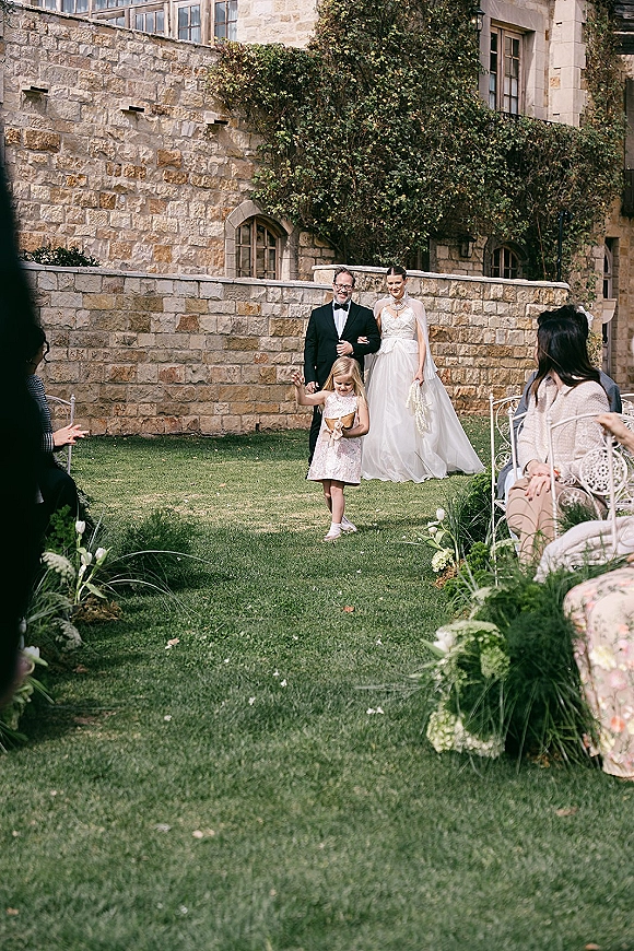 Wedding processional as bride walking down aisle with floral-lined greenery, escorted by father, flower girl with basket in ivy courtyard