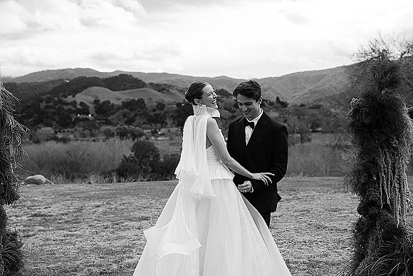 Couple portrait of bride laughing in a high-neck cape wedding dress with groom in black tuxedo beneath a floral arch in misty mountains