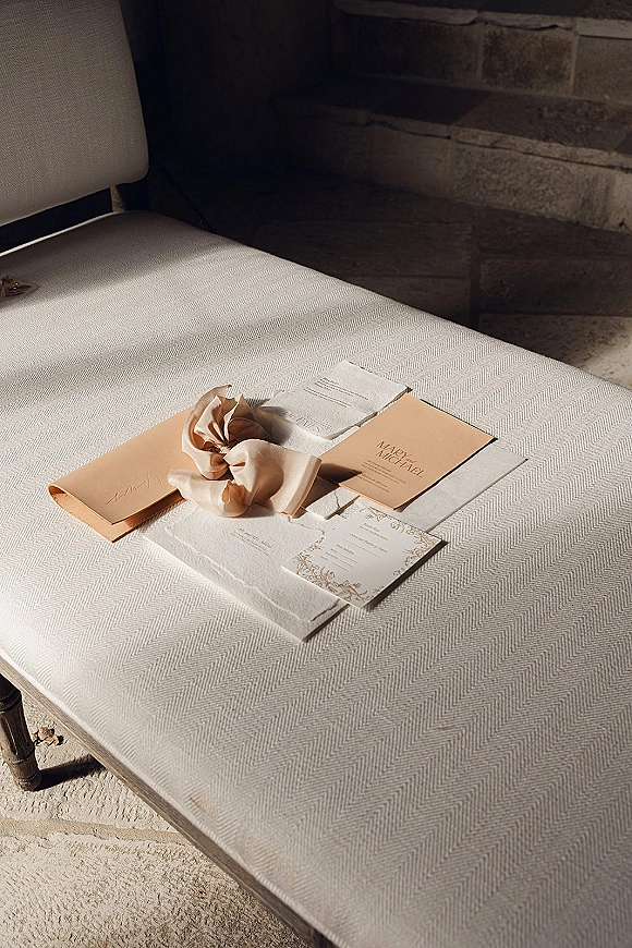 Invitation suite with neutral wedding invitations, envelopes and deckle-edge cards tied with a ribbon bow on a sunlit chair near stone steps