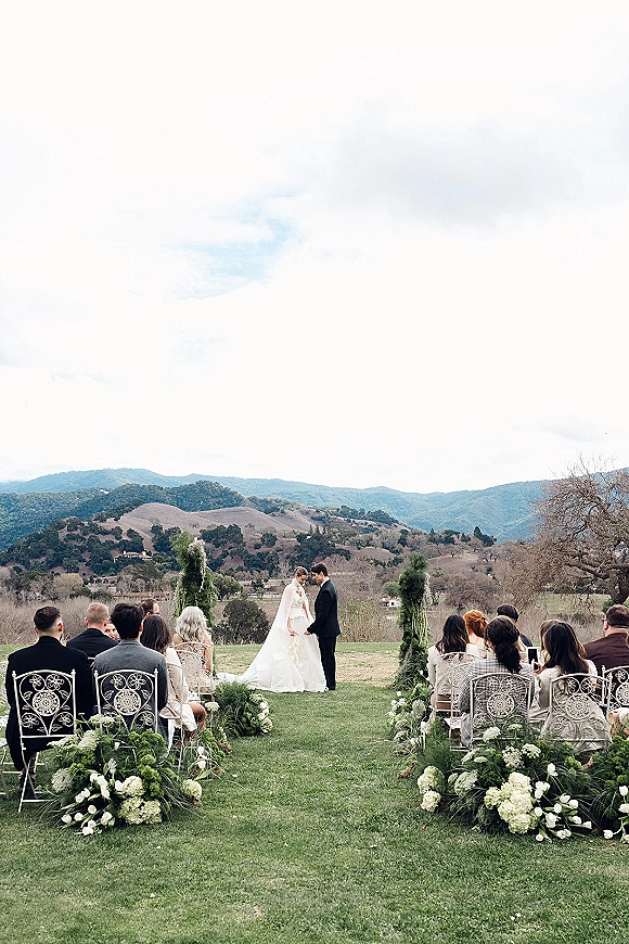 Wedding ceremony with bride and groom at altar, veil and tux, guests seated among white aisle florals on a lawn with mountain hills backdrop