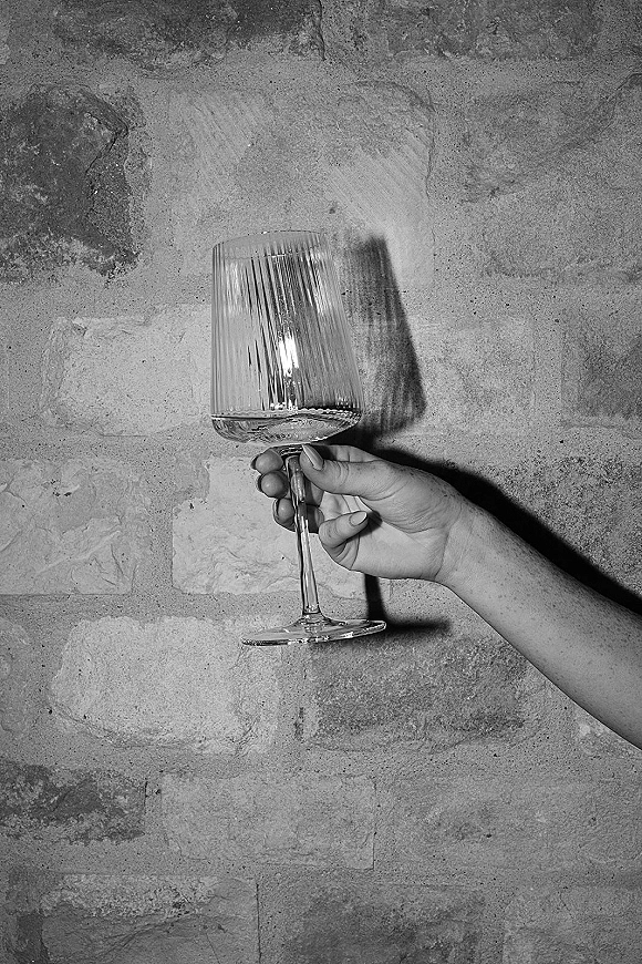 Wine glass toast held by a manicured hand with neutral nail polish, featuring a ribbed glass silhouette against a stone wall backdrop