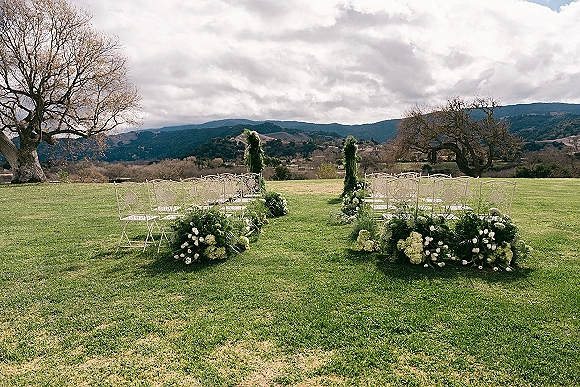 Ceremony aisle design with white chairs and hydrangea, tulip, and greenery garlands on a lawn with mountain views under clouds