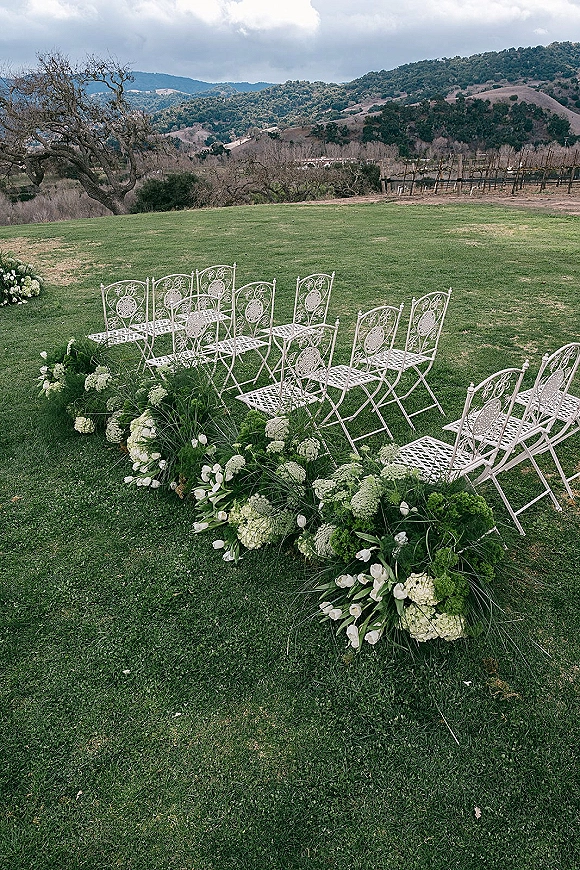 Ceremony aisle decor with outdoor ceremony aisle flowers lining white metal chairs, lush greenery and white blooms on a lawn with vineyard rows and mountains