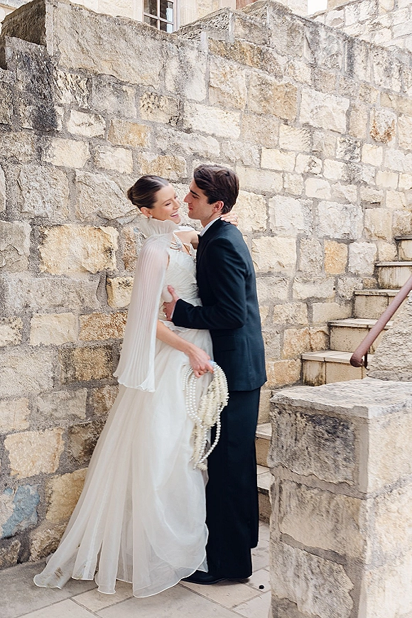 Couple portrait of bride and groom embrace, her cape veil and pearl strands over a high-neck gown beside a stone staircase wall