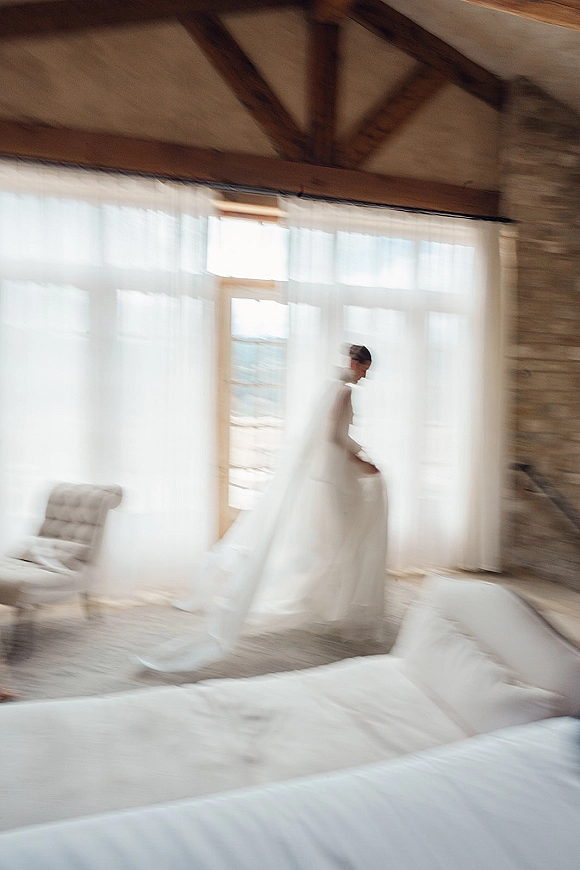 Bridal portrait of a bride in veil standing by a large window, backlit through sheer curtains, long veil flowing in a rustic suite