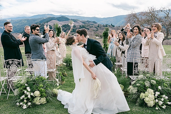 Wedding kiss as the groom dips the bride in her veil holding a bouquet, with white hydrangea aisle flowers and mountains behind