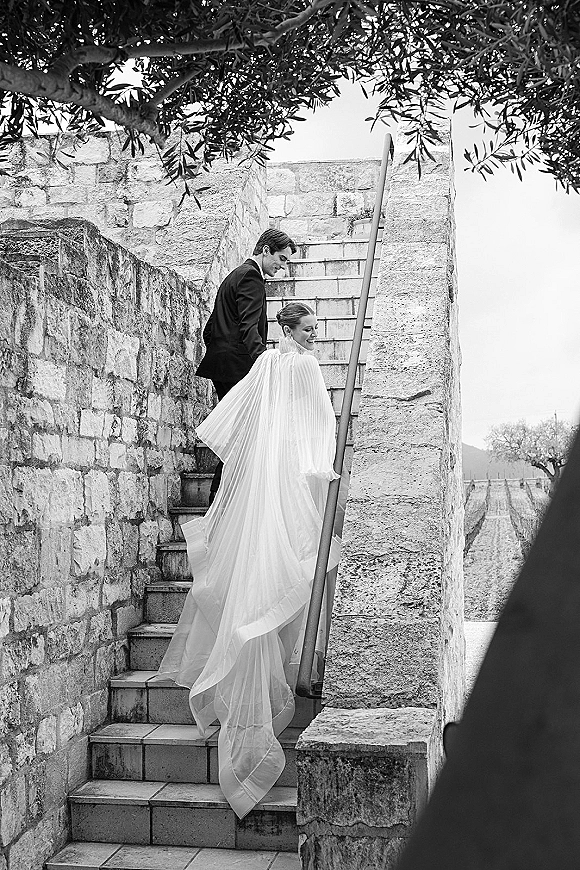 Couple portrait in black and white showing bride in cape veil and groom in tuxedo walking up stone stairs with vineyard behind