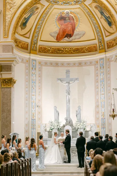 Ceremony moment as the bride and groom hold hands at a church wedding ceremony before a crucifix, flanked by light-blue bridesmaids.