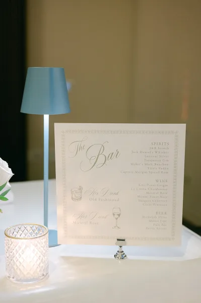 Wedding bar sign featuring a printed wedding bar menu, warmly lit by a table lamp beside it on the tabletop.