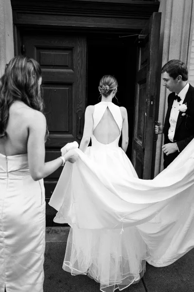 Ceremony entrance as the bride enters the venue, the groom holds the wooden door while a bridesmaid lifts the bridal train against a stone wall.