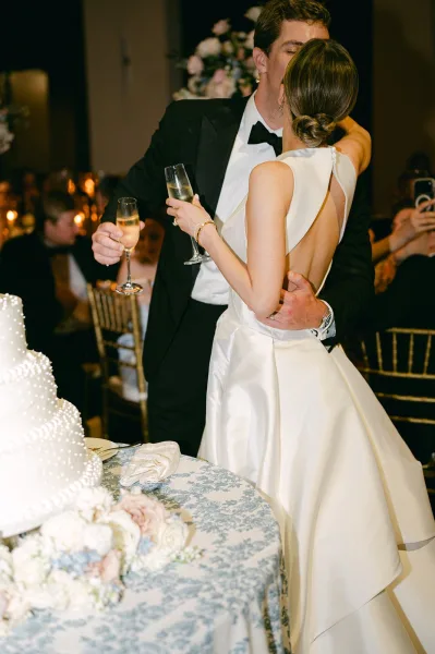 Wedding kiss portrait as the couple shares a champagne toast moment beside their wedding cake at a candlelit reception among guests.