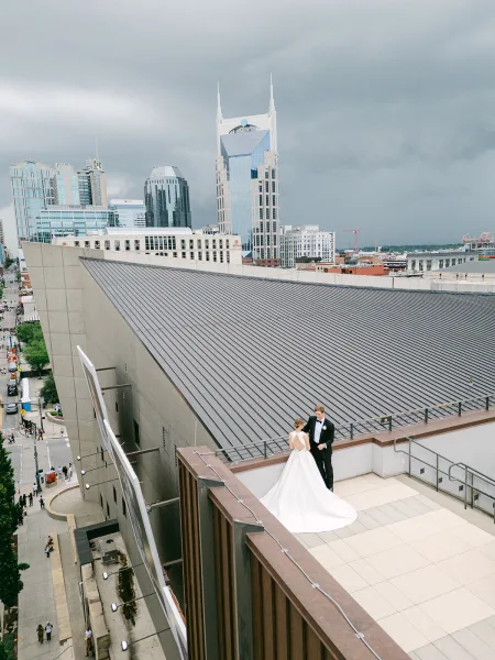 Rooftop couple portrait of newlyweds in a flowing bridal gown, a city skyline wedding photo against cloudy high-rises and street below.