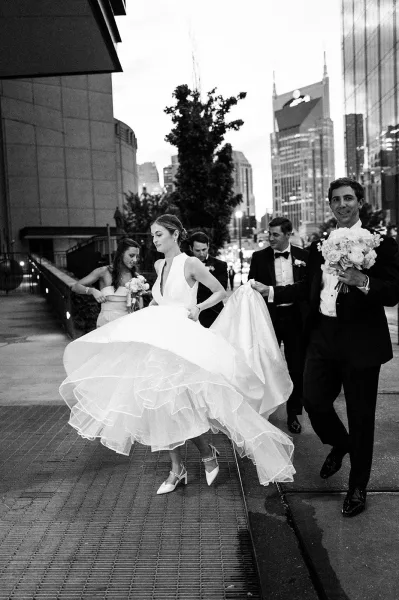 Bride and groom walking with their wedding party along a city sidewalk, her ball gown sweeping softly as the skyline rises behind them.