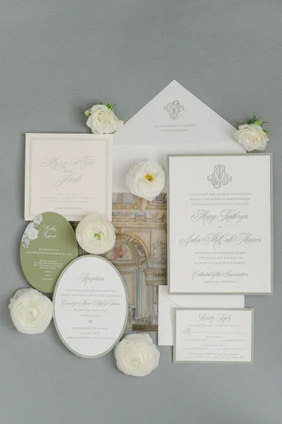 Wedding invitation suite with sage green wedding invitations and a monogram crest, styled on gray linen fabric.