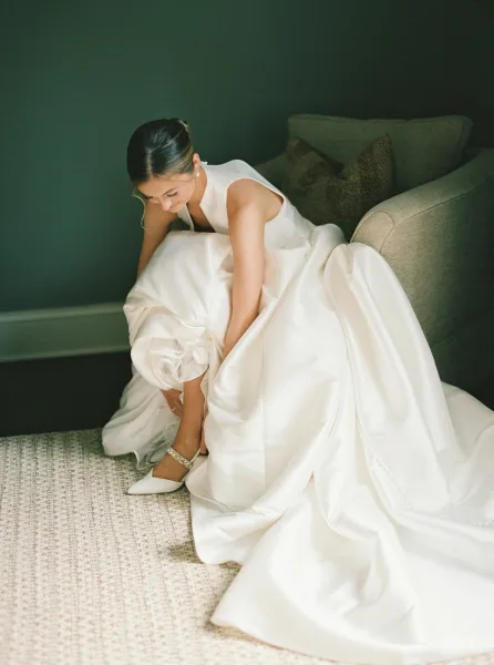 Bride getting ready, bride putting on shoes, pearl stud earrings catching light as she sits on a woven rug by a painted wall.
