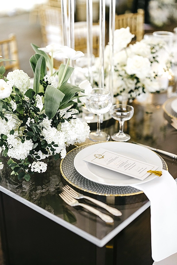 Reception tablescape with wedding table setting featuring white floral centerpiece, greenery, tall glass candle holders and place card on charger plate