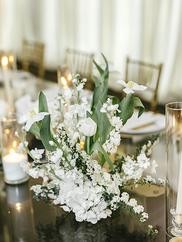 Wedding centerpiece with a white floral centerpiece in a glass vase, floating candles and greenery on a candlelit reception table with white draping