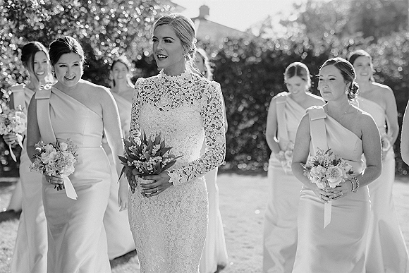 Bride with bridesmaids walking with bride in a sunlit garden, all holding matching bouquets as trees blur softly behind them