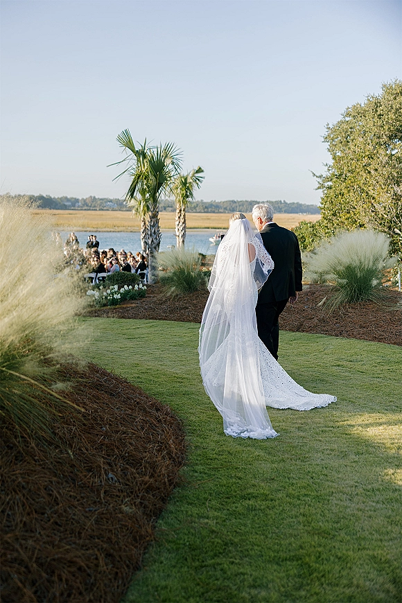 Bride walking down aisle with father in tuxedo, lace gown and cathedral veil flowing behind, past seated guests on a palm-lined lawn by marsh water.