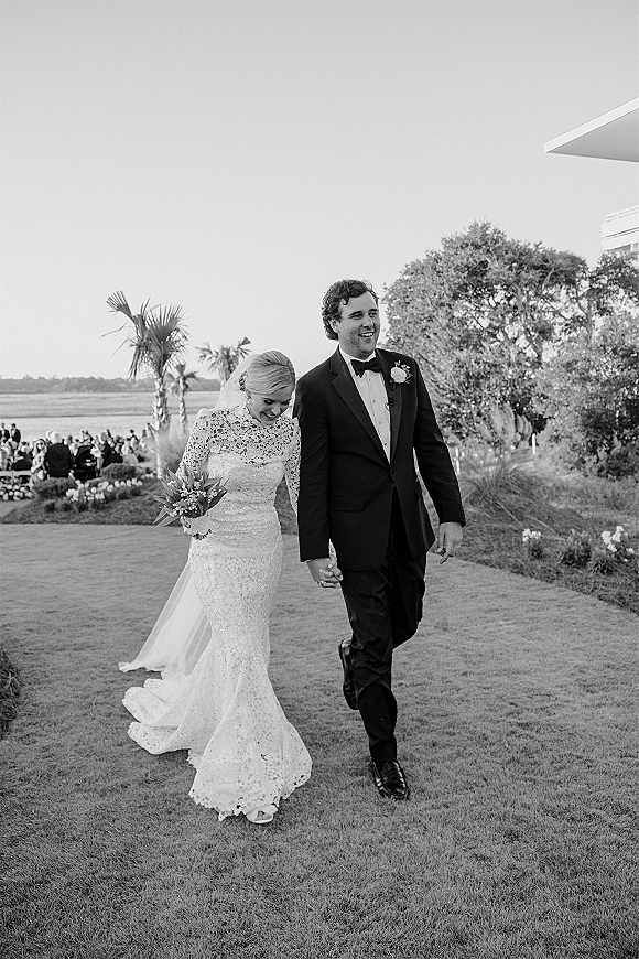 Wedding recessional as bride and groom walk the aisle holding hands, bride in lace long sleeves with veil and bouquet, guests by waterfront lawn