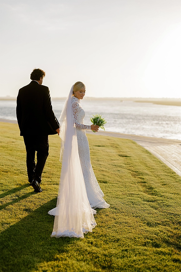 Couple portrait of bride and groom walking, her cathedral veil and bouquet trailing on an oceanfront boardwalk at sunset