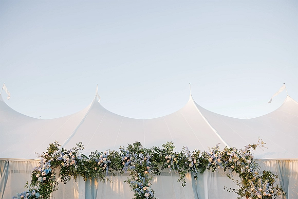 Wedding tent decor with a floral garland of greenery and pastel blooms draped over a white sailcloth entrance against blue sky