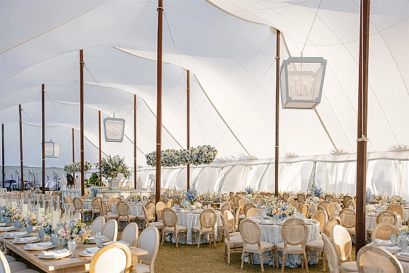 Tent wedding reception under a white sailcloth tent with hanging lantern chandeliers, round tables in blue linens, and rattan chairs on lawn