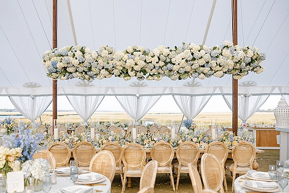 Reception tablescape with wedding tent reception styling, blue-and-white hydrangeas overhead, candles and rattan chairs in a sailcloth tent by the marsh view