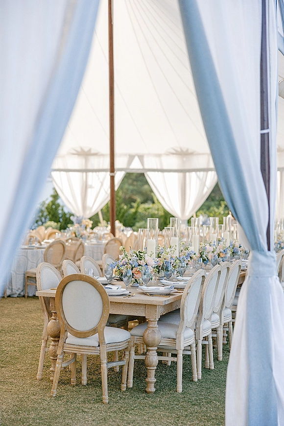 Reception tablescape at an outdoor tent reception with long wood farm tables, pastel florals, glass cylinder vases, and white taper candles on a lawn