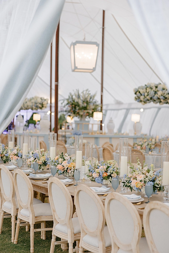 Reception tablescape with a long banquet table wedding setup, floral garland and candles under a clear tent with string lights on lawn