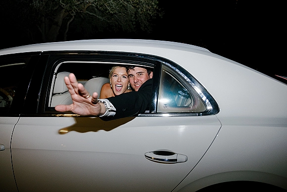 Wedding send off as newlyweds in car wave from a white limousine at night, bride in wedding dress beside groom in tuxedo under trees