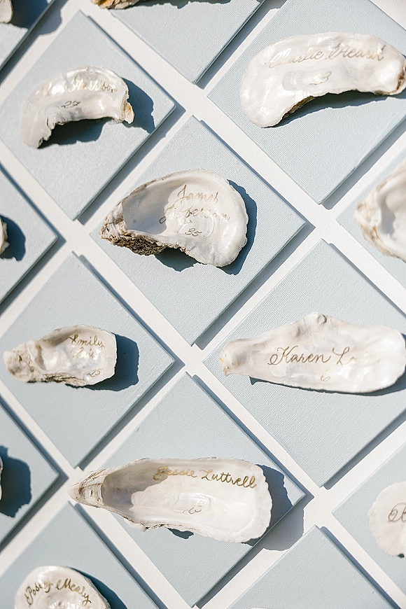 Oyster escort cards on oyster shells with gold calligraphy, arranged over light blue paper cards on a clean white surface flat lay