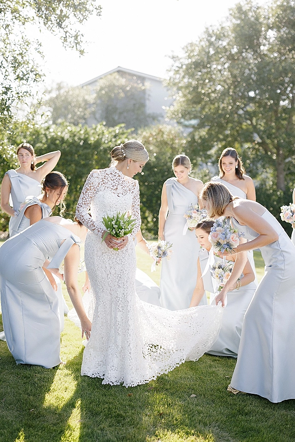 Bride with bridesmaids fixing dress train, her long sleeve lace wedding dress and bouquet glowing on a sunny lawn with trees and a house