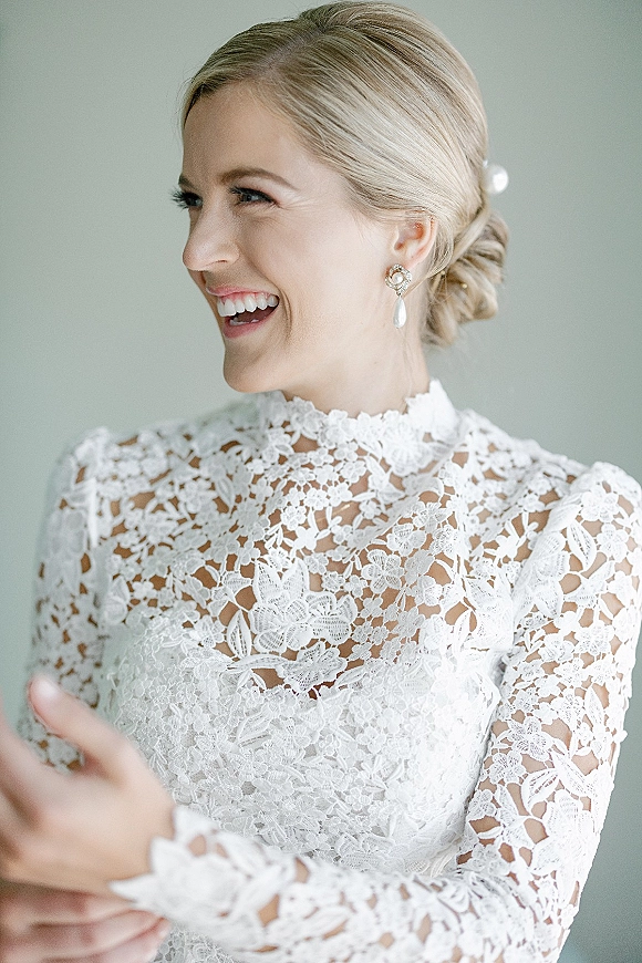 Bridal portrait of a bride laughing in side profile, wearing a high neck lace wedding dress and pearl drop earrings by a neutral wall
