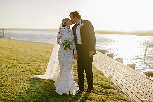 Wedding kiss as bride and groom kiss on a waterfront dock at sunset, her lace long sleeve dress and veil trailing, bouquet in hand