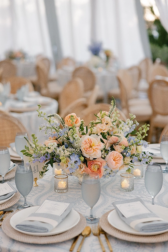 Reception tablescape with wedding floral centerpiece of peach roses and blue blooms, gold flatware, blue goblets, and votive candles in a white tent