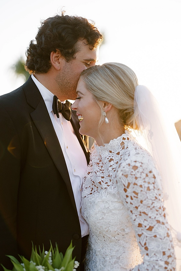 Wedding couple portrait with groom kissing bride’s forehead at sunset, bride in lace dress and veil holding greenery bouquet in soft backlight