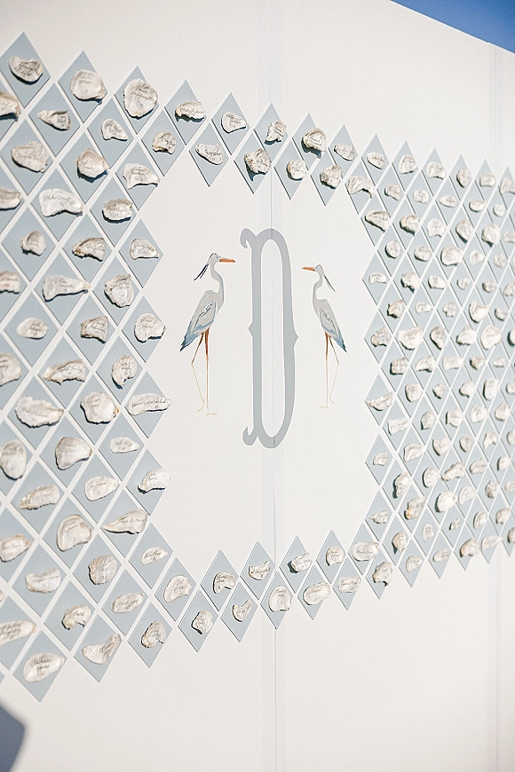 Seating chart display with oyster shell seating chart escort cards in calligraphy on a diamond grid, featuring a monogram and painted birds on a white wall