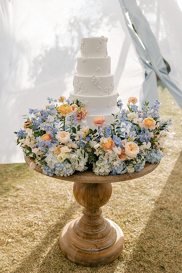 Wedding cake four tier wedding cake with white fondant and floral appliqués, topped with pastel blooms on a wooden pedestal by draped fabric outdoors