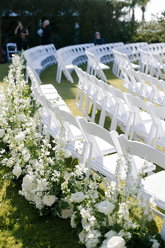 Ceremony aisle decor with white folding chairs and white rose florals lining a greenery garland runner on a garden lawn with hedges
