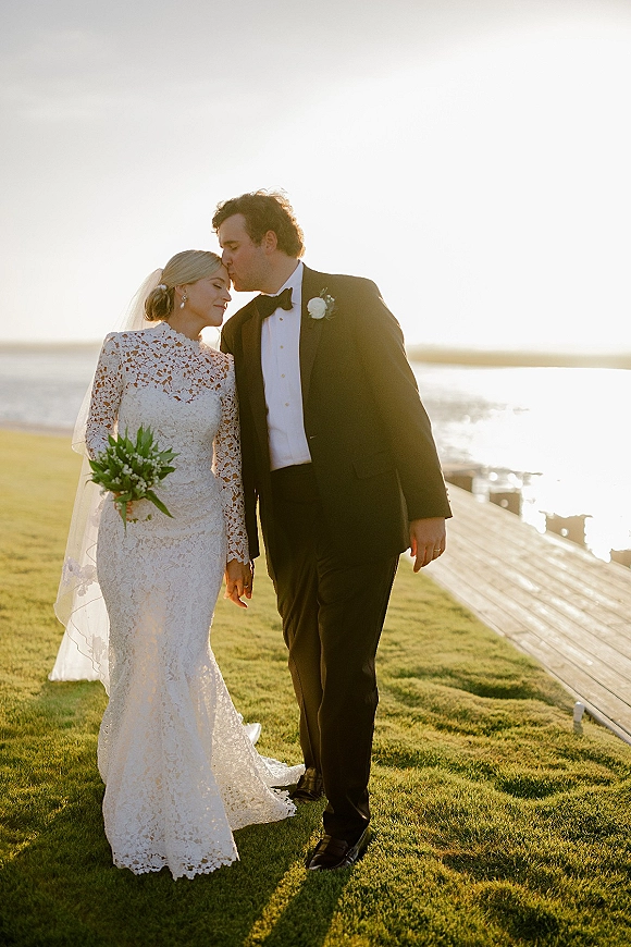 Couple portrait of bride and groom sharing a wedding forehead kiss, her veil and bouquet glowing by the ocean at sunset on a boardwalk