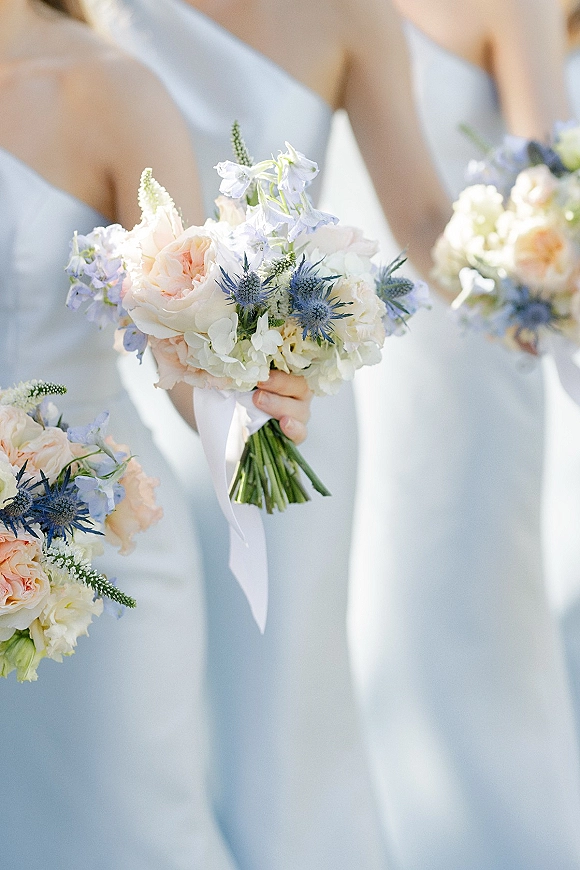 Bridesmaid bouquets with a peach and blue bouquet mix of roses, blue thistle, and greenery tied with white ribbon in soft outdoor light