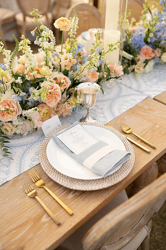 Reception tablescape with wedding table centerpiece of pastel roses and blue blooms, pillar candles, gold flatware on a wood table outdoors