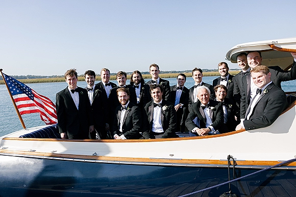 Groomsmen group photo on a yacht, black tuxedos with bow ties and boutonnières, with an American flag over blue water and marsh shoreline